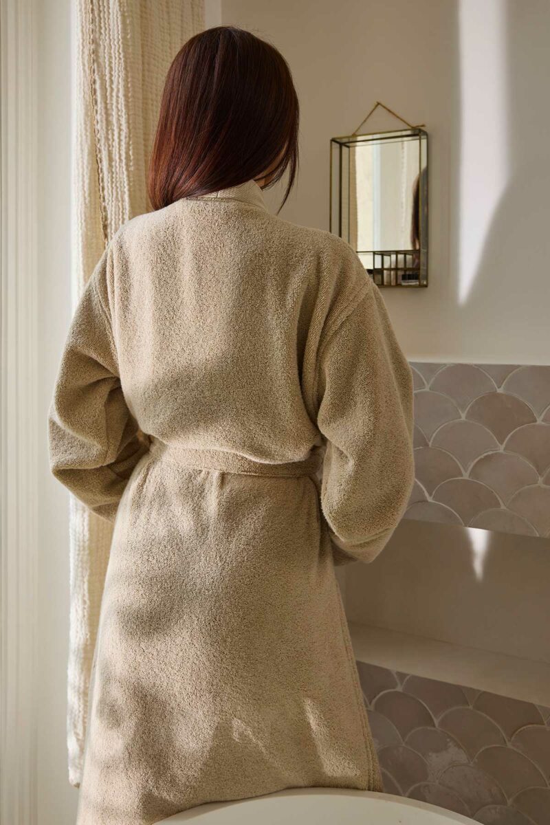 Femme de dos portant le peignoir kimono Narumi Madura en beige sable dans une salle de bains lumineuse