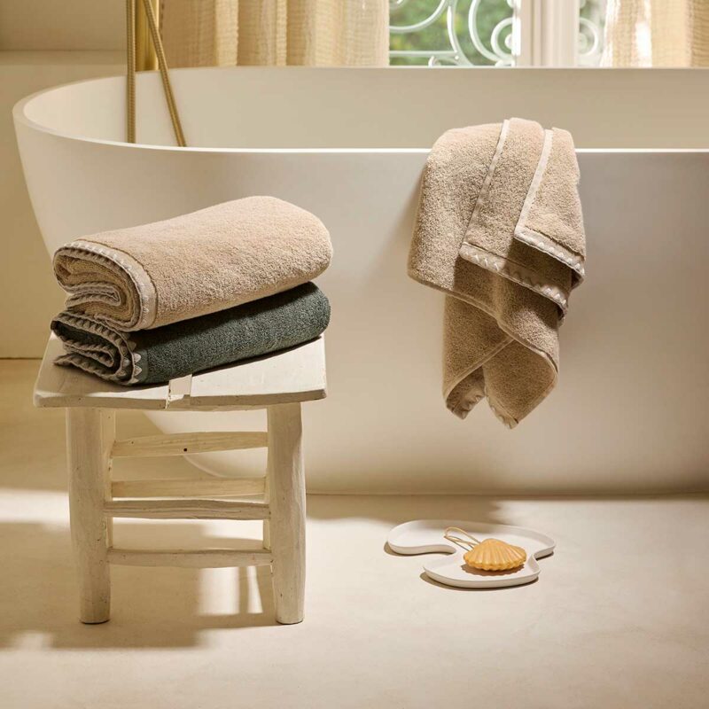 Serviettes de bain Neryda Madura en beige et vert séquoia posées sur un tabouret devant une baignoire îlot